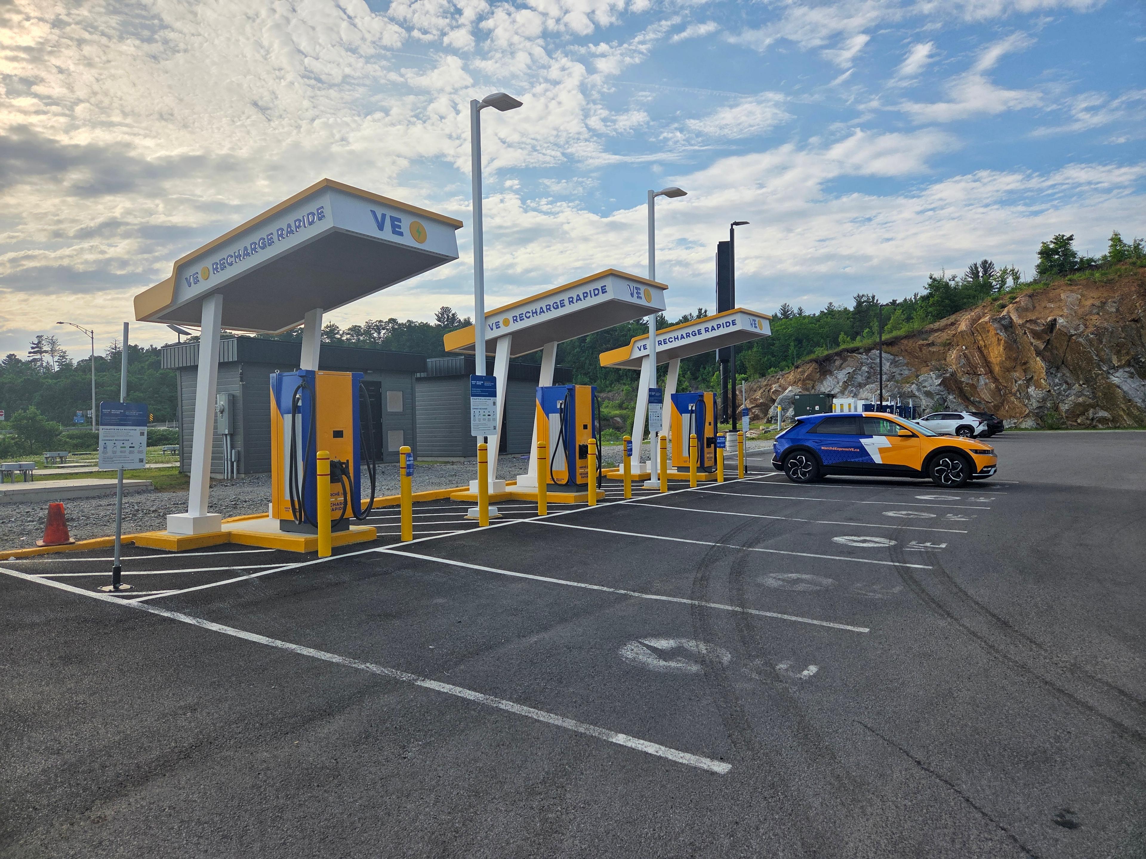 The photo shows a VE Recharge Rapide electric vehicle charging station with yellow and blue chargers. A blue and yellow EV with a large white lightning bolt graphic and MarcheExpressVE.ca branding is parked and plugged in.
